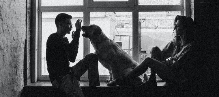 grayscale-of-man-woman-and-dog-on-window-2173360